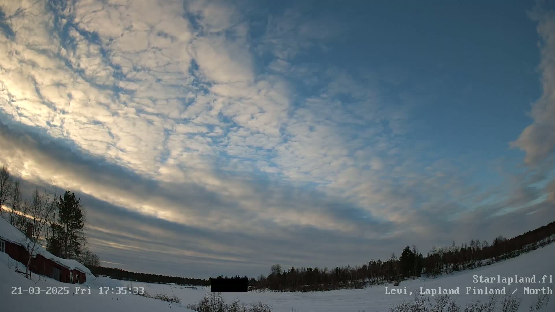 A mostly clear, blue sky with thin, scattered clouds covers a snowy, wooded landscape at dusk, featuring a red building to the left and a dark, rectangular object in the middle distance.