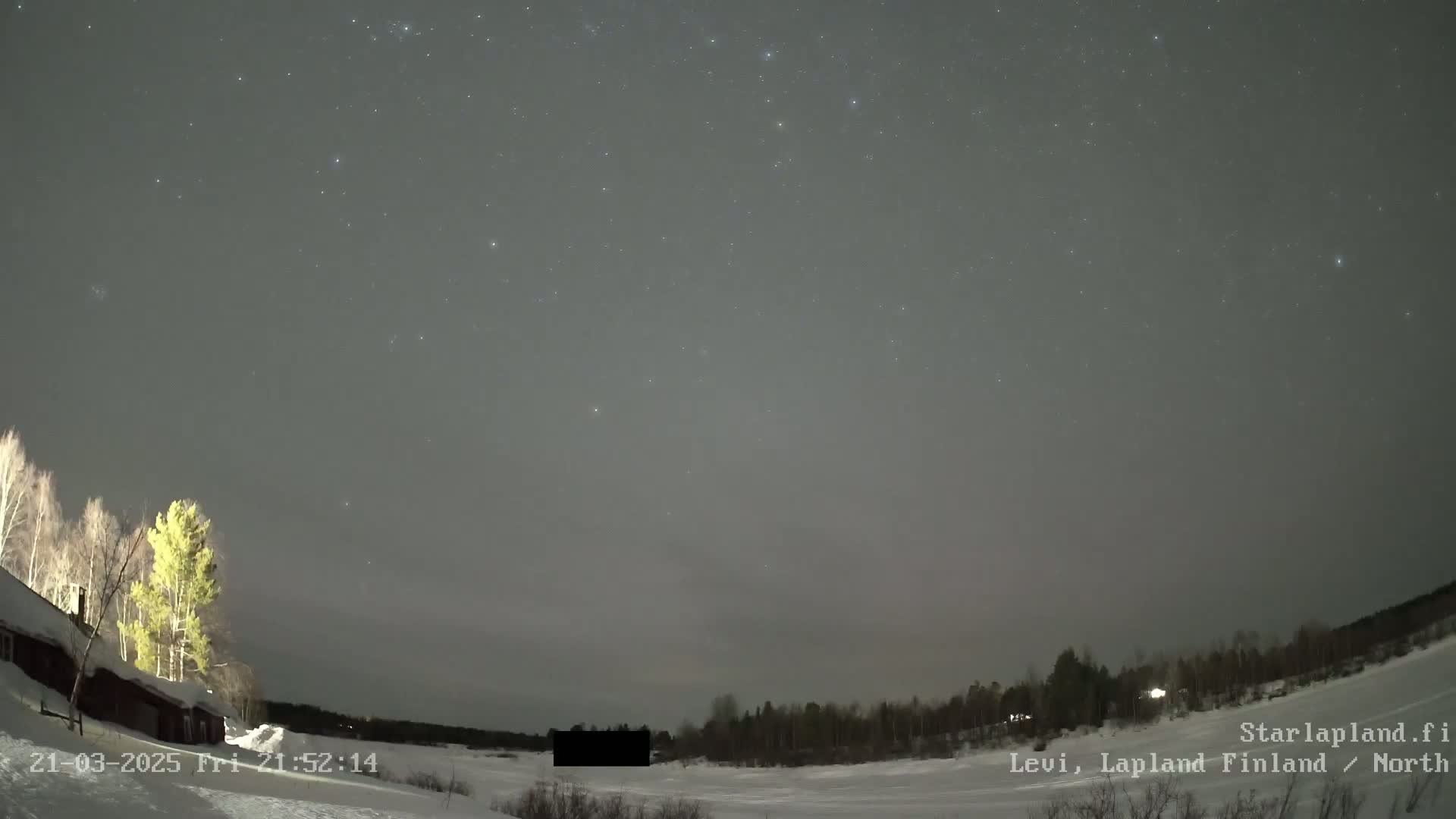 A nighttime, snowy landscape with a mostly clear sky dotted with stars is visible above a dark building and a line of trees.