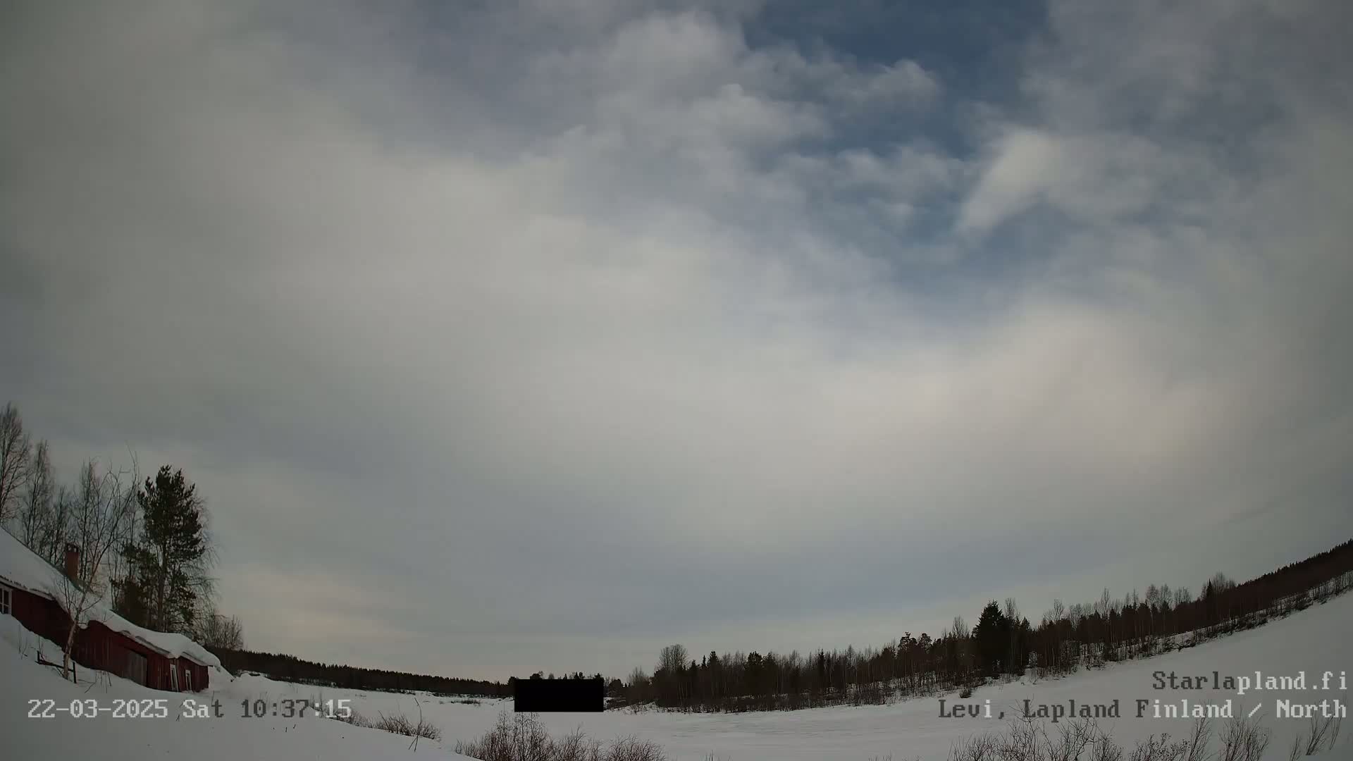 A mostly cloudy, overcast sky over a snow-covered landscape with a red building and a line of dark trees.