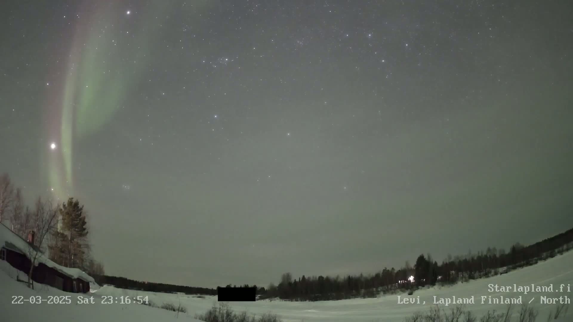 A vibrant green and pink aurora borealis illuminates a night sky dotted with stars over a snow-covered landscape featuring a dark structure and a line of trees.