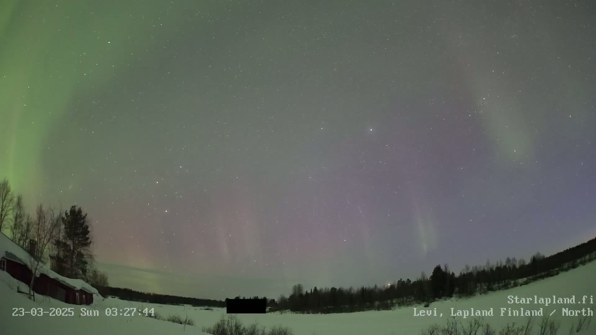 A vibrant aurora borealis illuminates a night sky above a snow-covered landscape with dark trees and a small red building.