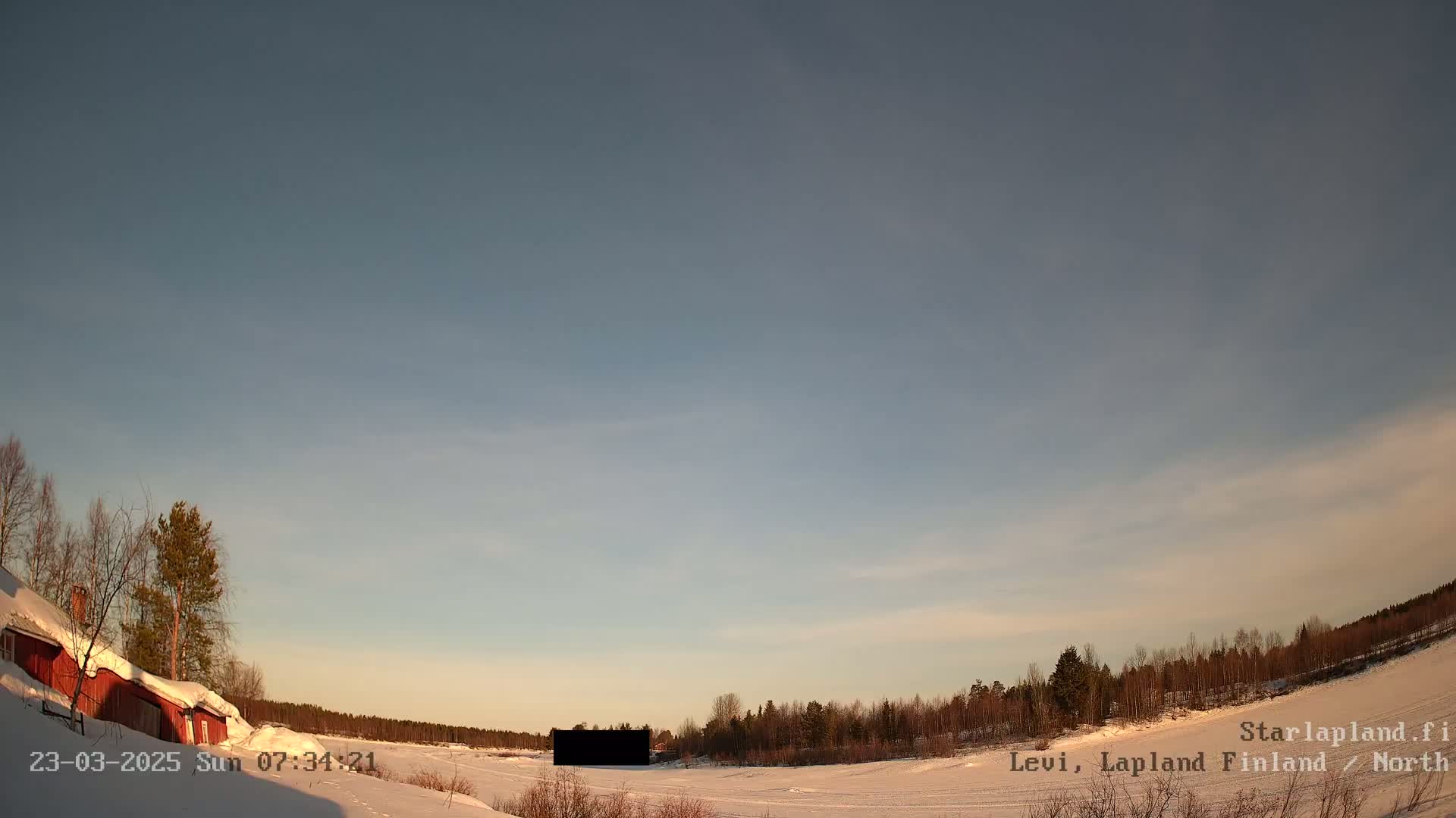 A snow-covered landscape under a mostly clear, pale blue sky shows a red building to the left and a dark rectangular object in the middle distance, with a line of leafless trees in the background.