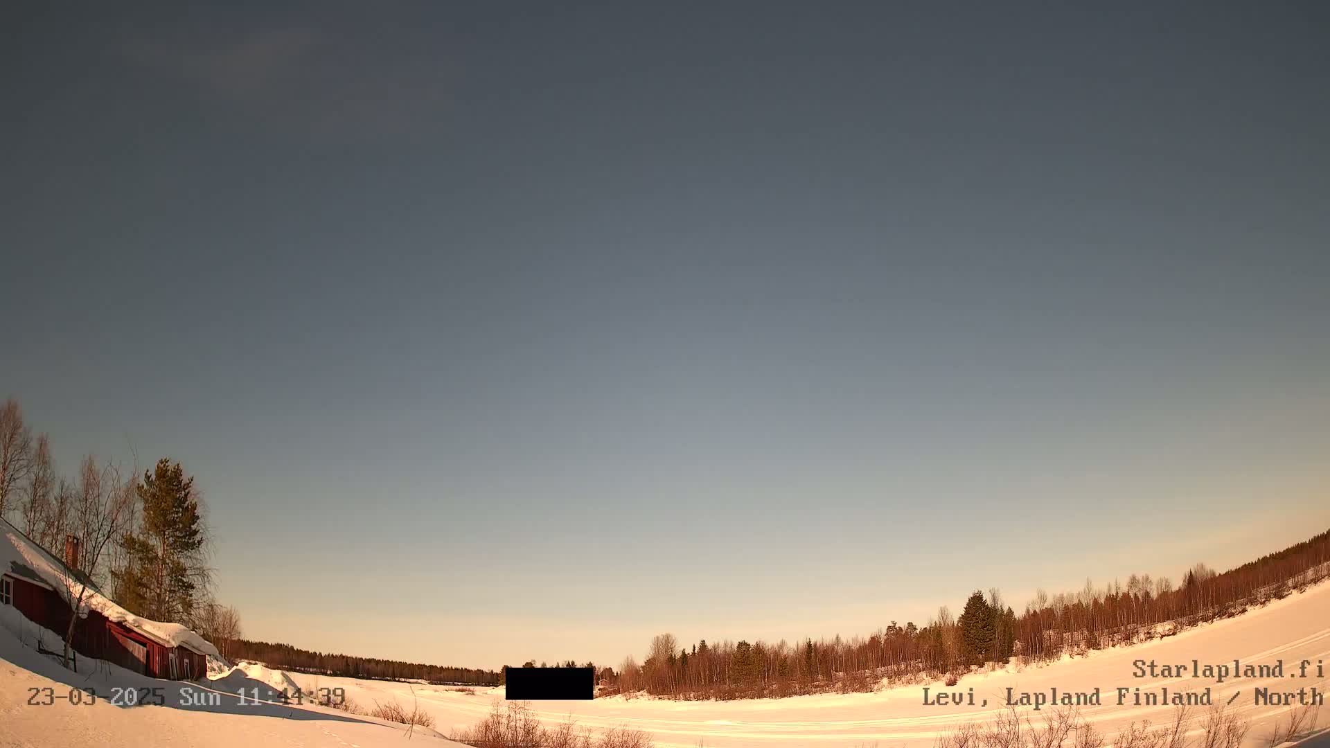 A snow-covered landscape with a red building and a line of trees under a clear, pale sky.