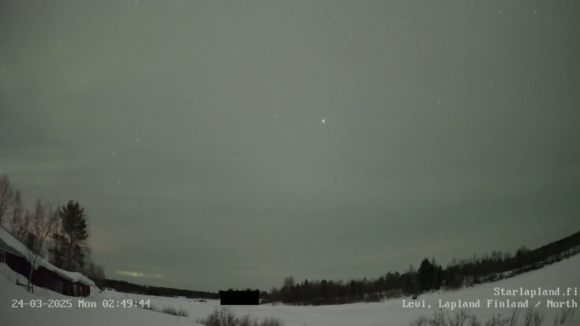 A faint aurora borealis illuminates a night sky over a snow-covered landscape with sparse, dark trees and a building.