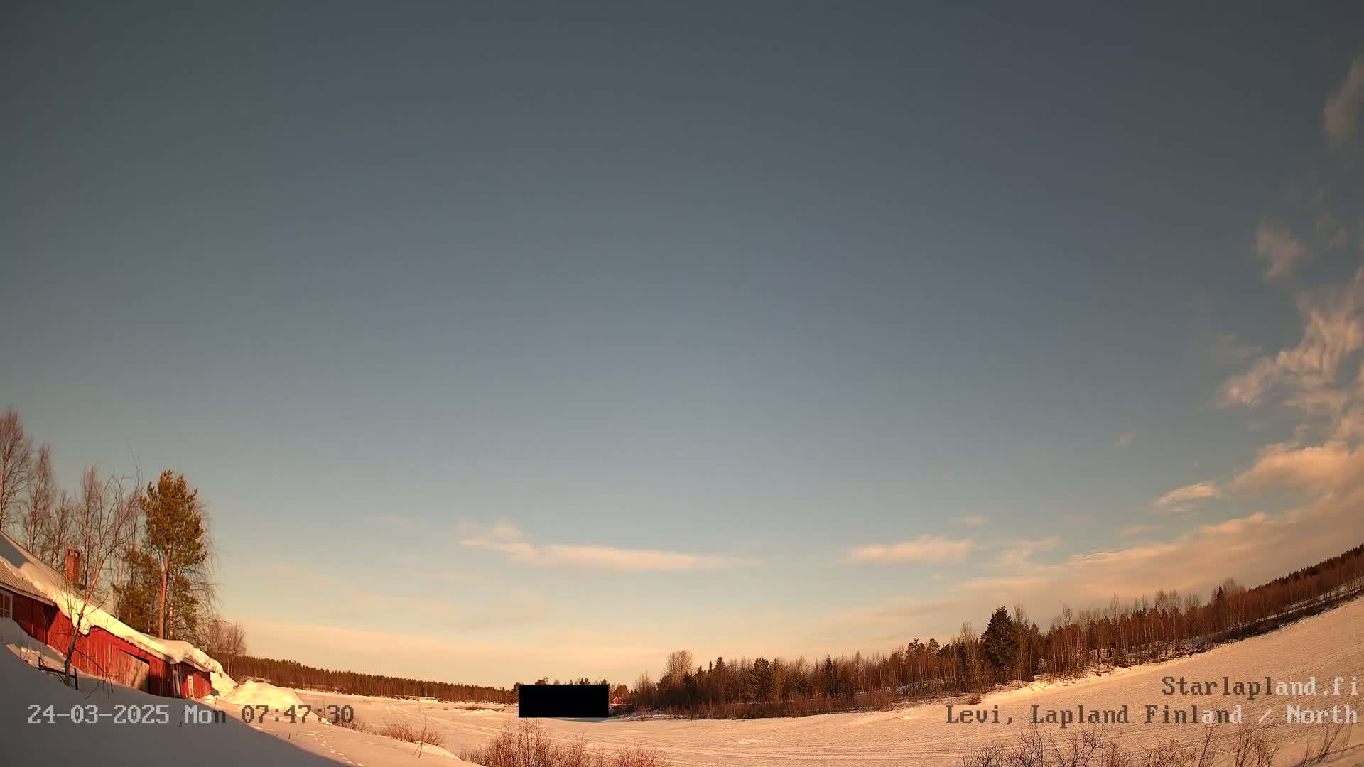A snow-covered landscape with a red building and a line of trees under a mostly clear, pale blue sky.