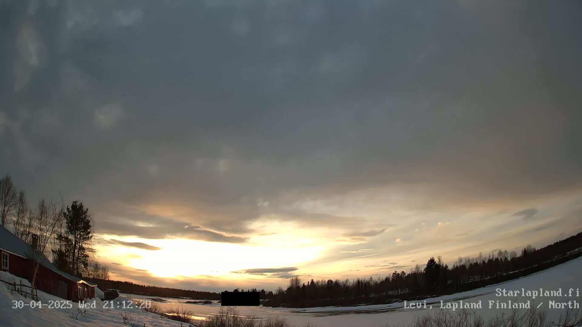 A partly cloudy sunset illuminates a snow-covered landscape with a red building and dark trees in the foreground.