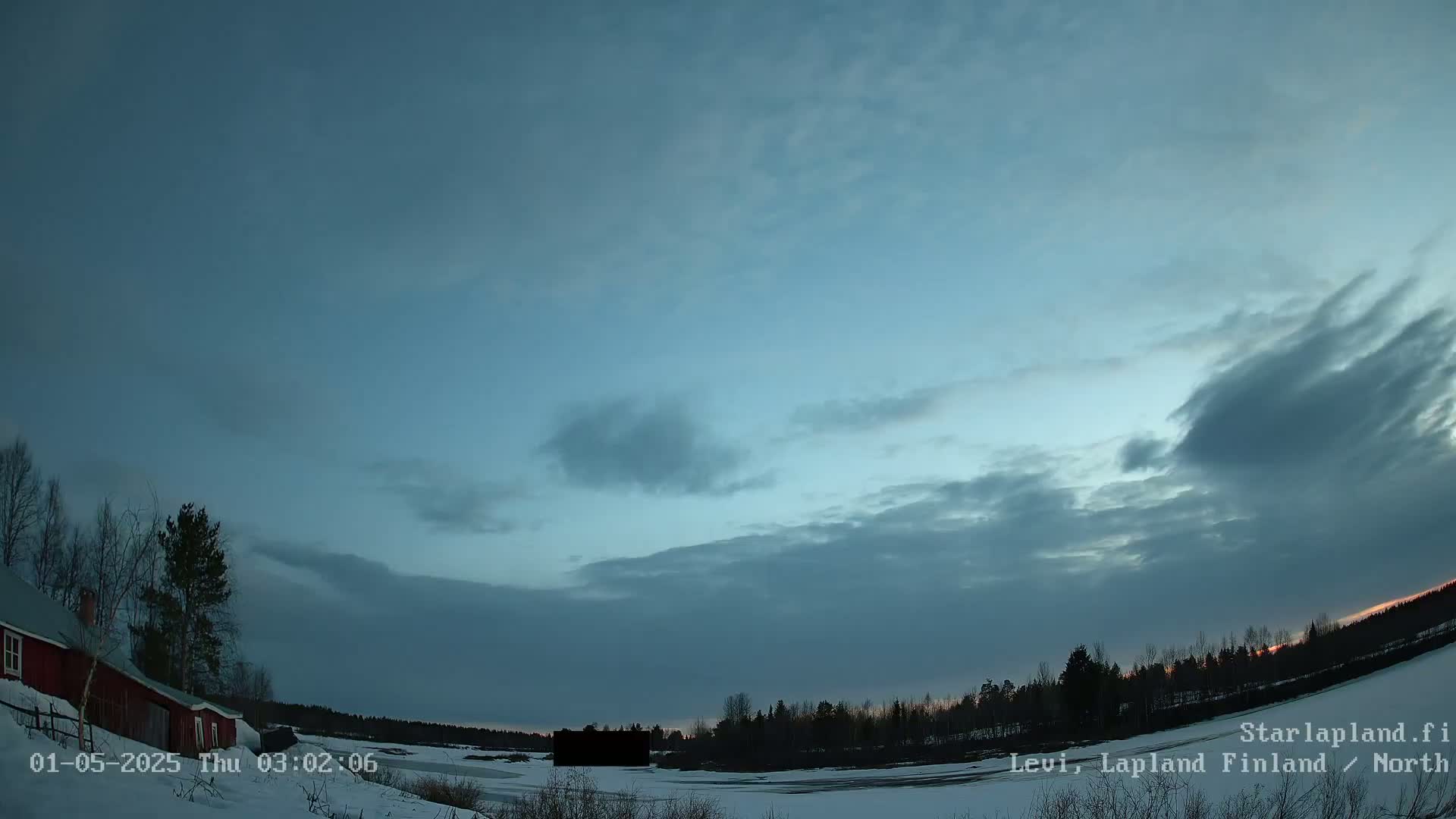 A partly cloudy twilight sky over a snow-covered landscape with a red building and dark trees visible near a frozen body of water.