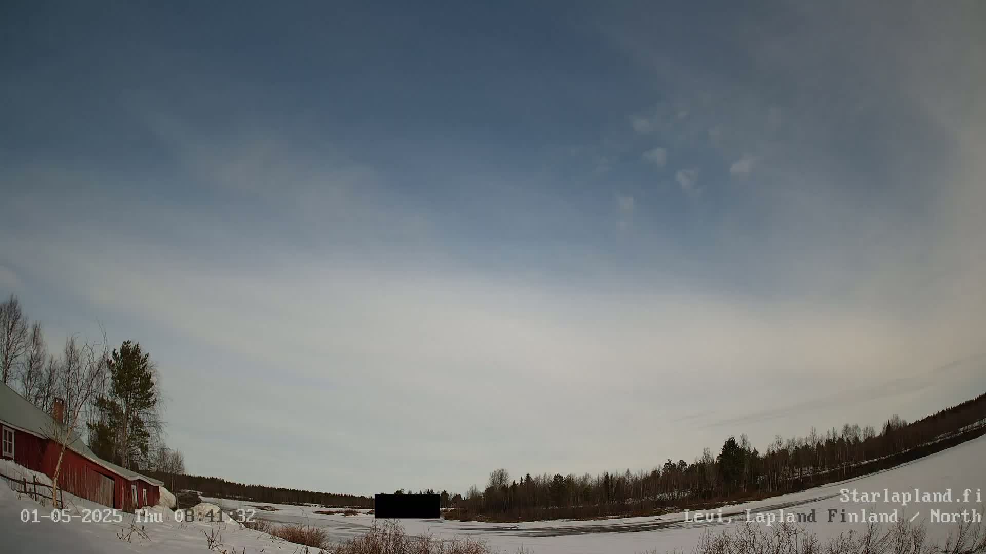 A red building sits on a snow-covered landscape next to a frozen body of water under a mostly clear, pale blue sky with a few wispy clouds.