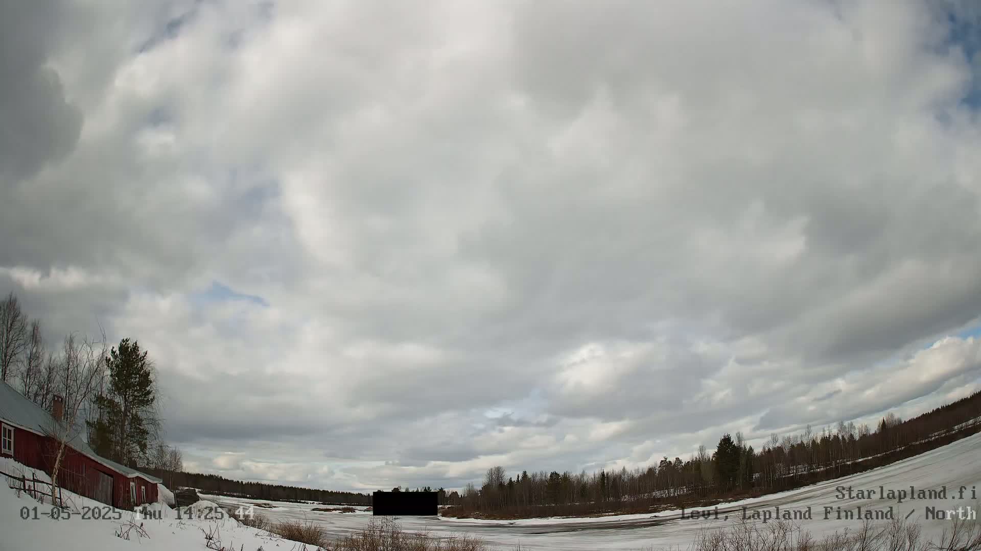 A partially frozen body of water is visible under a mostly cloudy sky, with a red building and bare trees to the left and a dark object in the mid-ground.