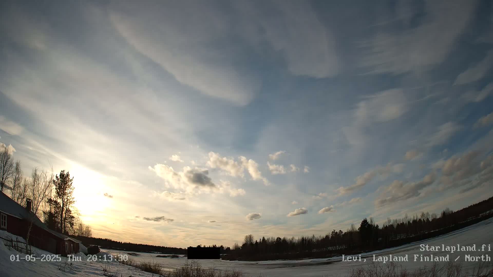 A mostly cloudy sunset over a snowy landscape with a few trees and a building visible near the bottom left.