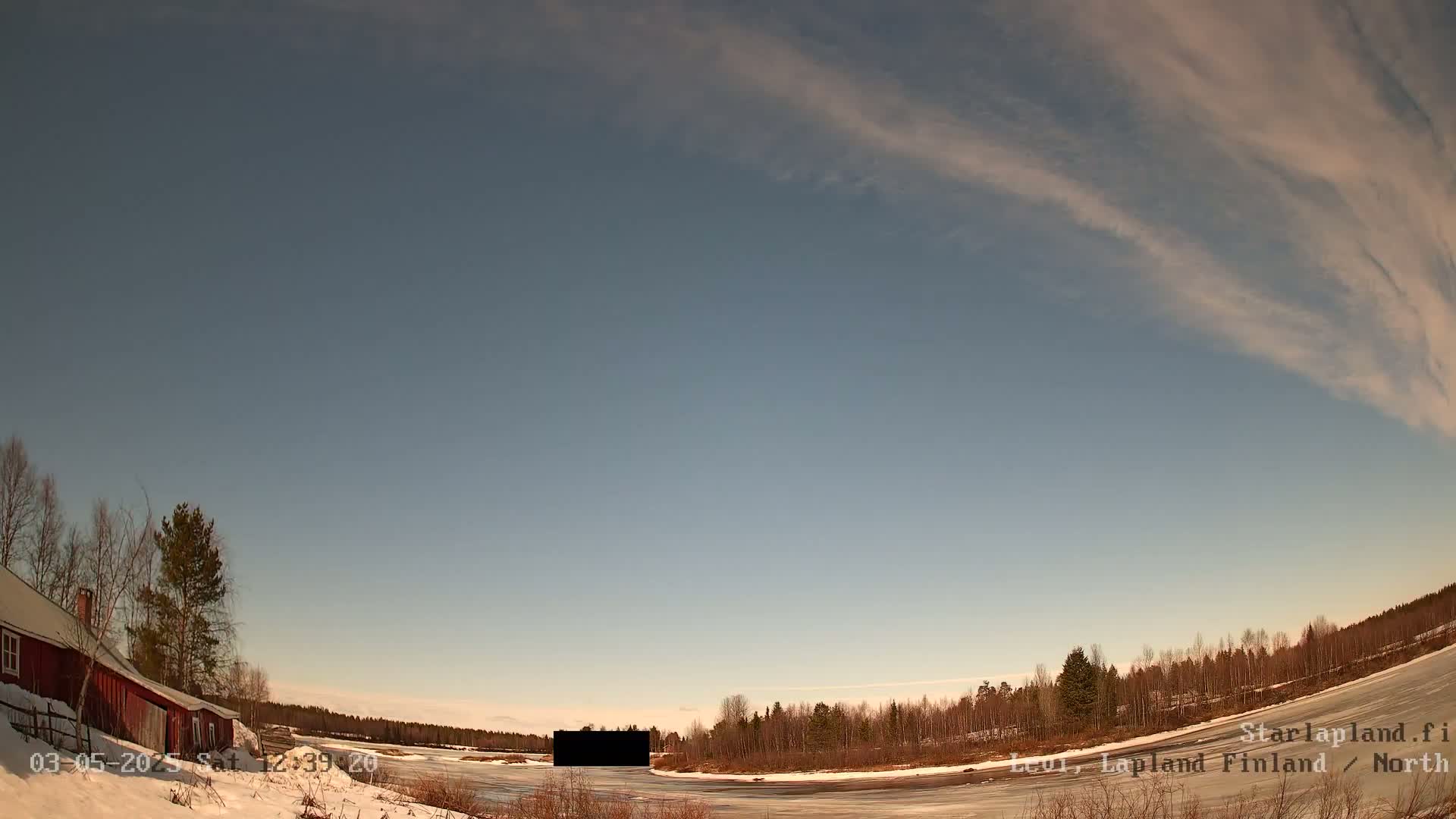 A red building sits near a snow-covered frozen body of water, with leafless trees lining the far shore under a mostly clear, light blue sky with wispy clouds.