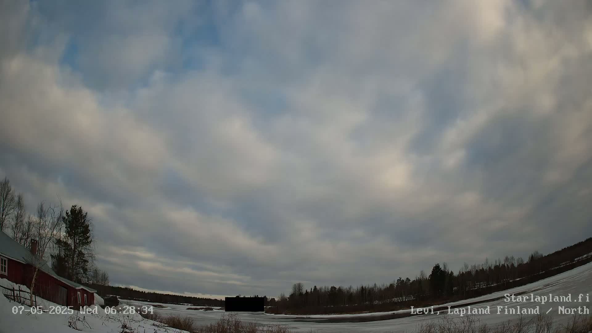 A mostly cloudy sky over a snow-covered landscape with a red building and a dark, rectangular object near a frozen body of water, bordered by a line of trees.