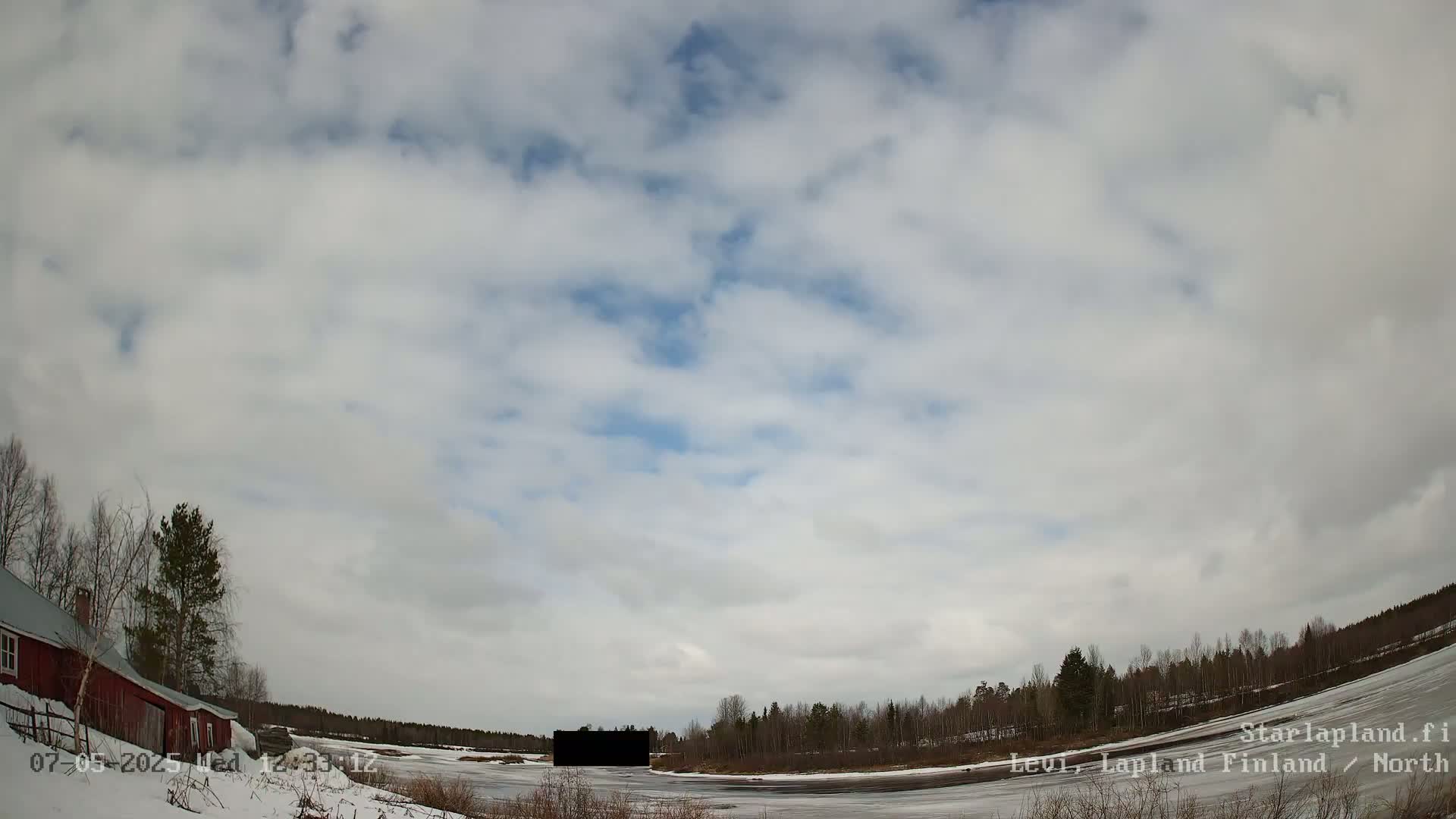 A partly cloudy sky over a snow-covered landscape with a frozen body of water, a red building, and leafless trees.
