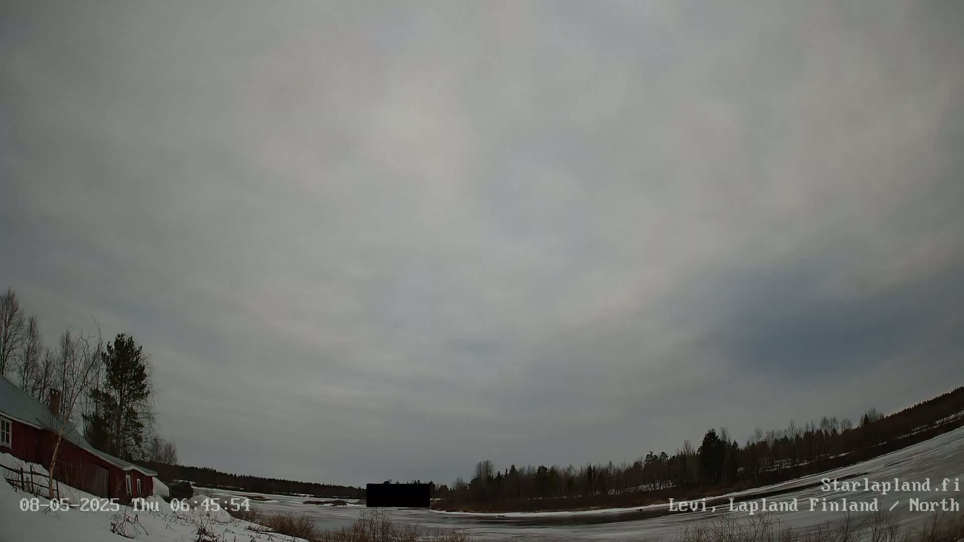 A mostly cloudy sky hangs over a snow-covered landscape featuring a red building, leafless trees, and a partially frozen body of water.
