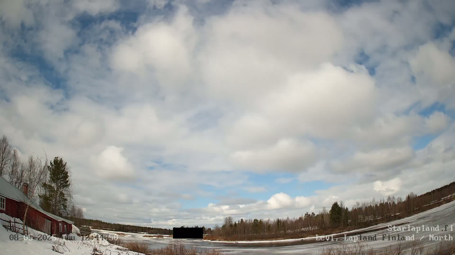 A partly cloudy sky over a snow-covered landscape with a red barn, trees, and a frozen body of water.