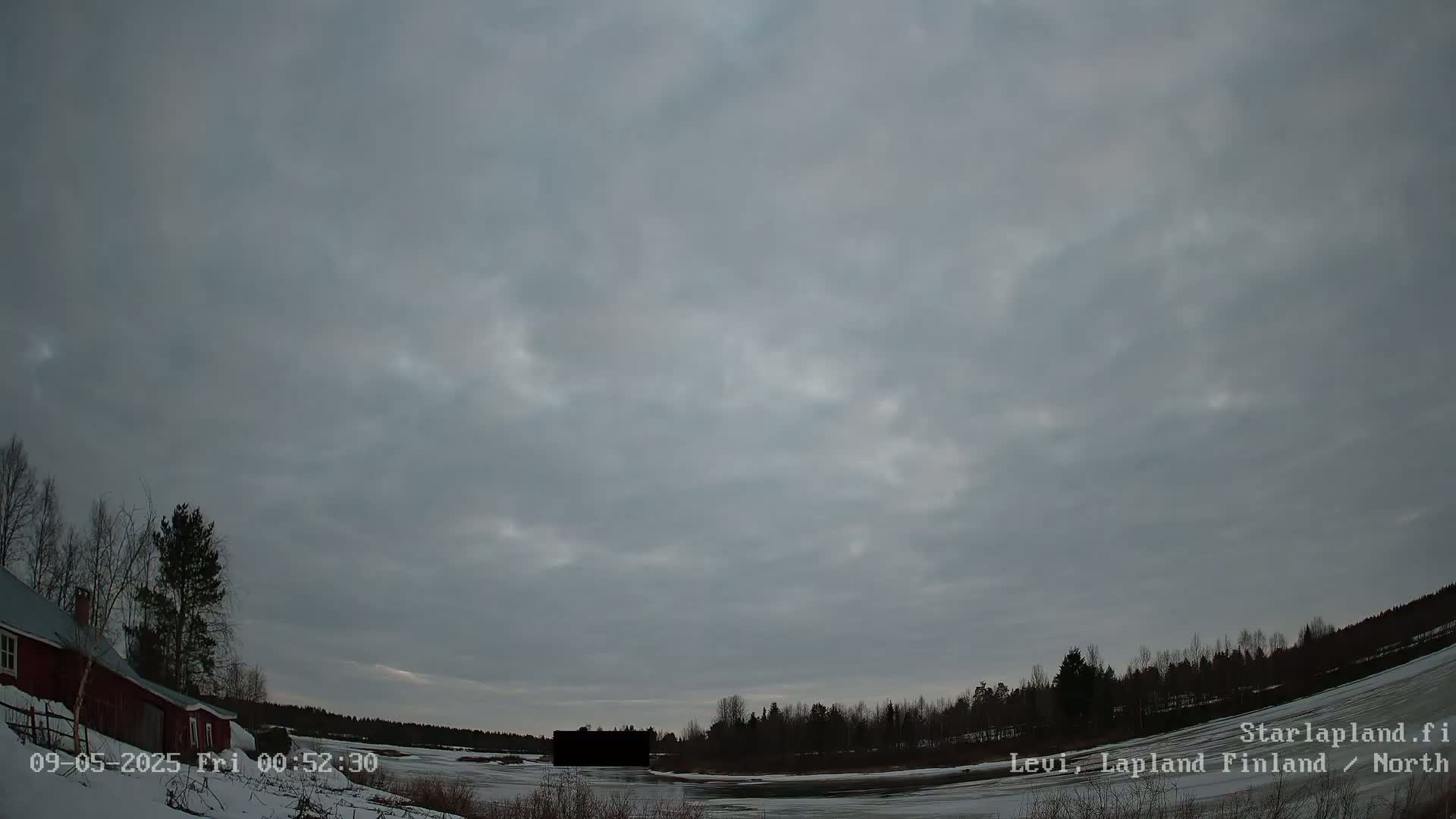 A mostly cloudy sky hangs over a snow-covered landscape featuring a red building, bare trees, and a partially frozen body of water.