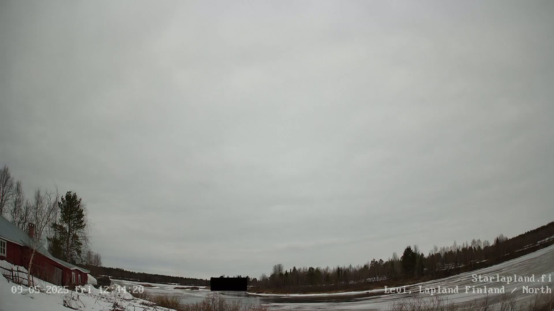 A red building sits near a snow-covered riverbank under an overcast sky.