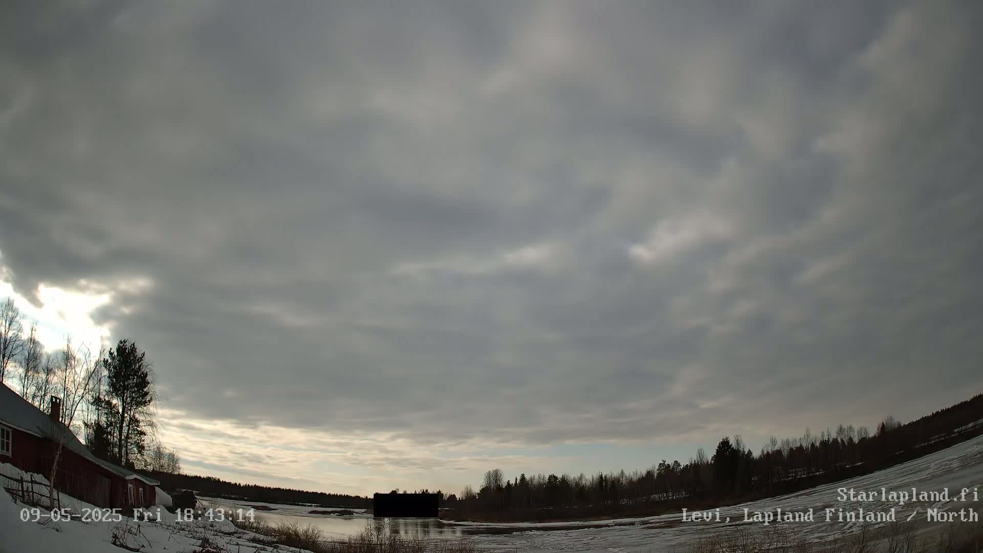 A mostly cloudy sky over a snow-covered landscape with a red building, some trees, and a body of water in the distance.