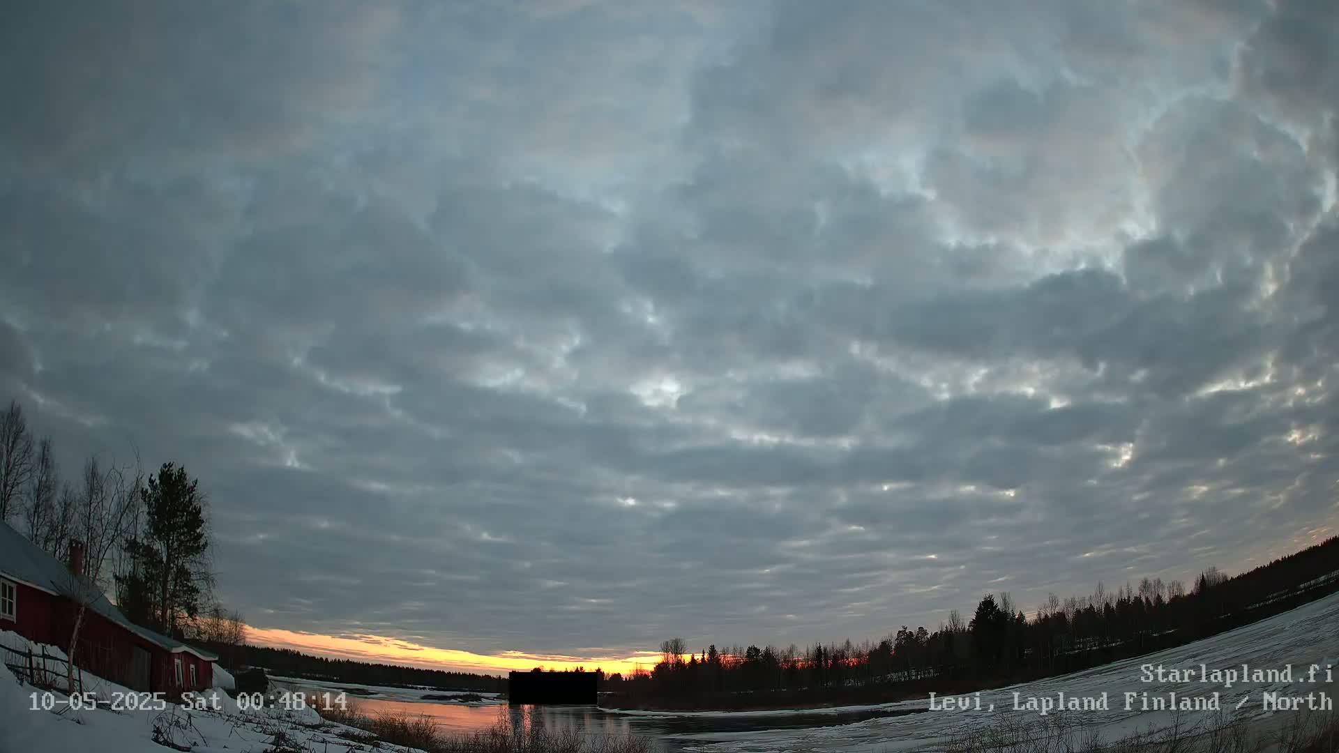 A partially cloudy sunset over a snow-covered landscape with a red house and a body of water in the foreground.