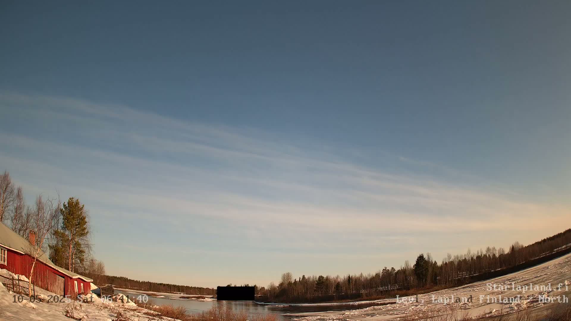 A red building sits on a snow-covered bank overlooking a partially frozen river under a mostly clear, pale blue sky with thin, wispy clouds.