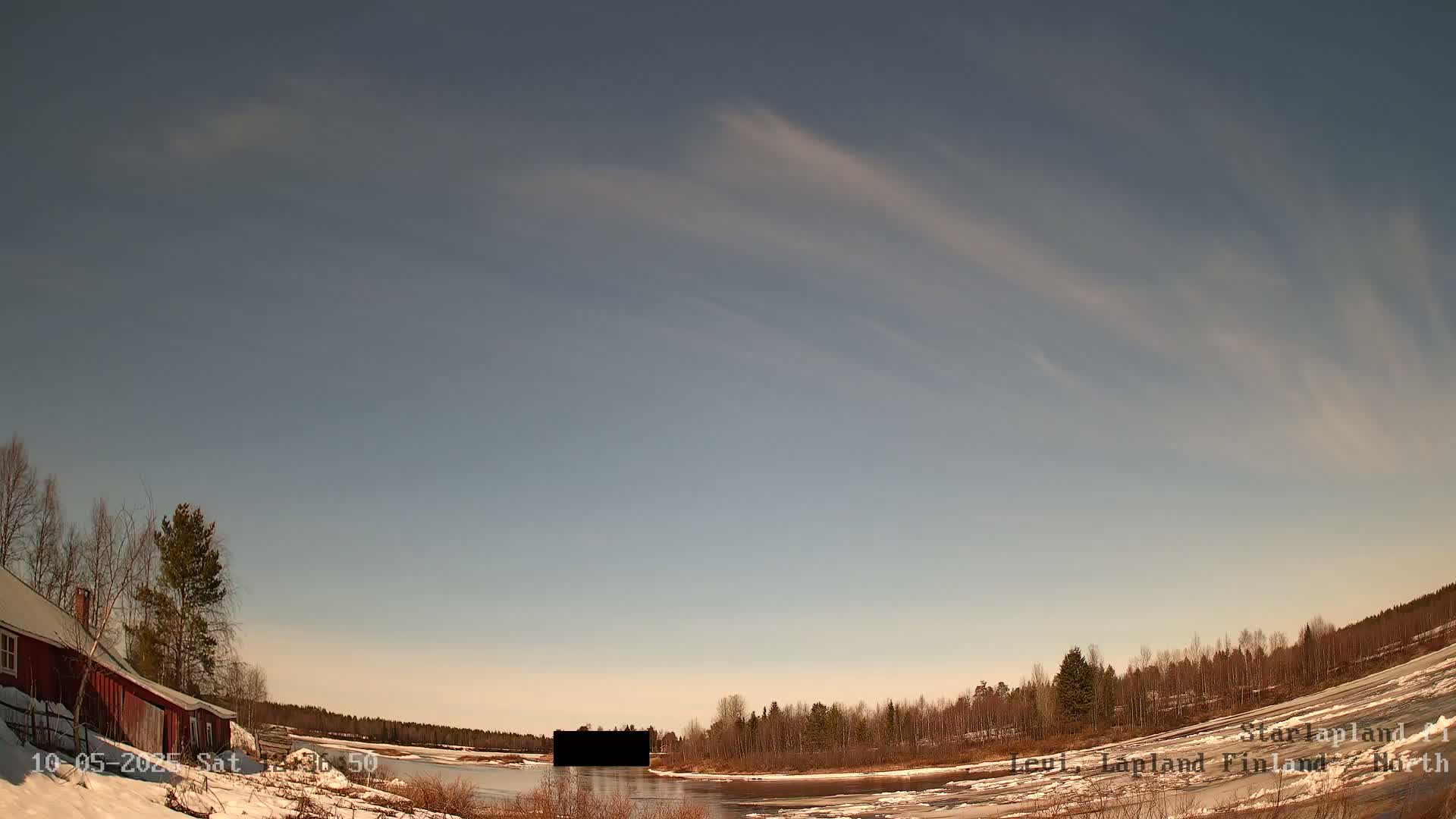 A partly sunny day with wispy clouds over a snow-covered landscape featuring a red building near a partially frozen river and a line of trees.