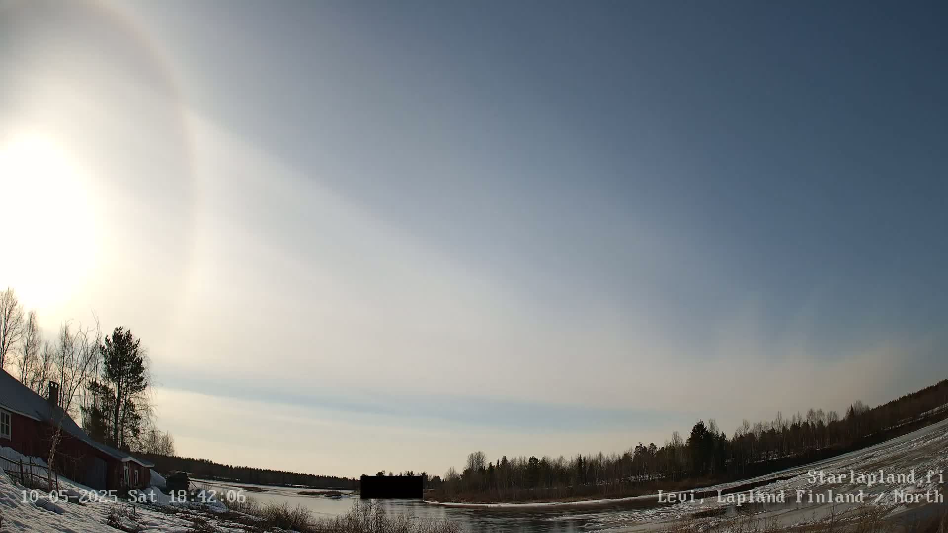 A bright, partly cloudy day with a 22° halo around the sun shows a snow-covered landscape with a red building and dark trees near a partially frozen river.