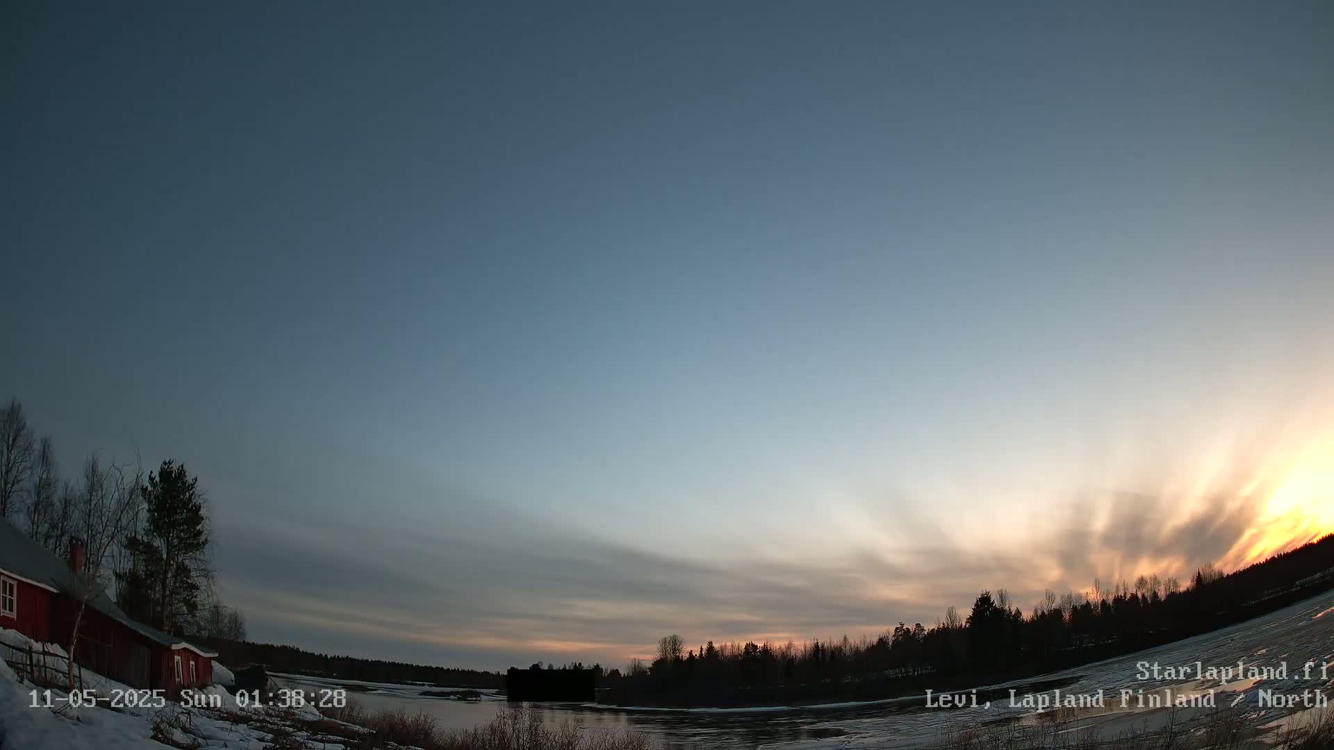 A red cabin sits on a snow-covered shore beside a frozen lake under a twilight sky with streaks of orange and yellow near the horizon.