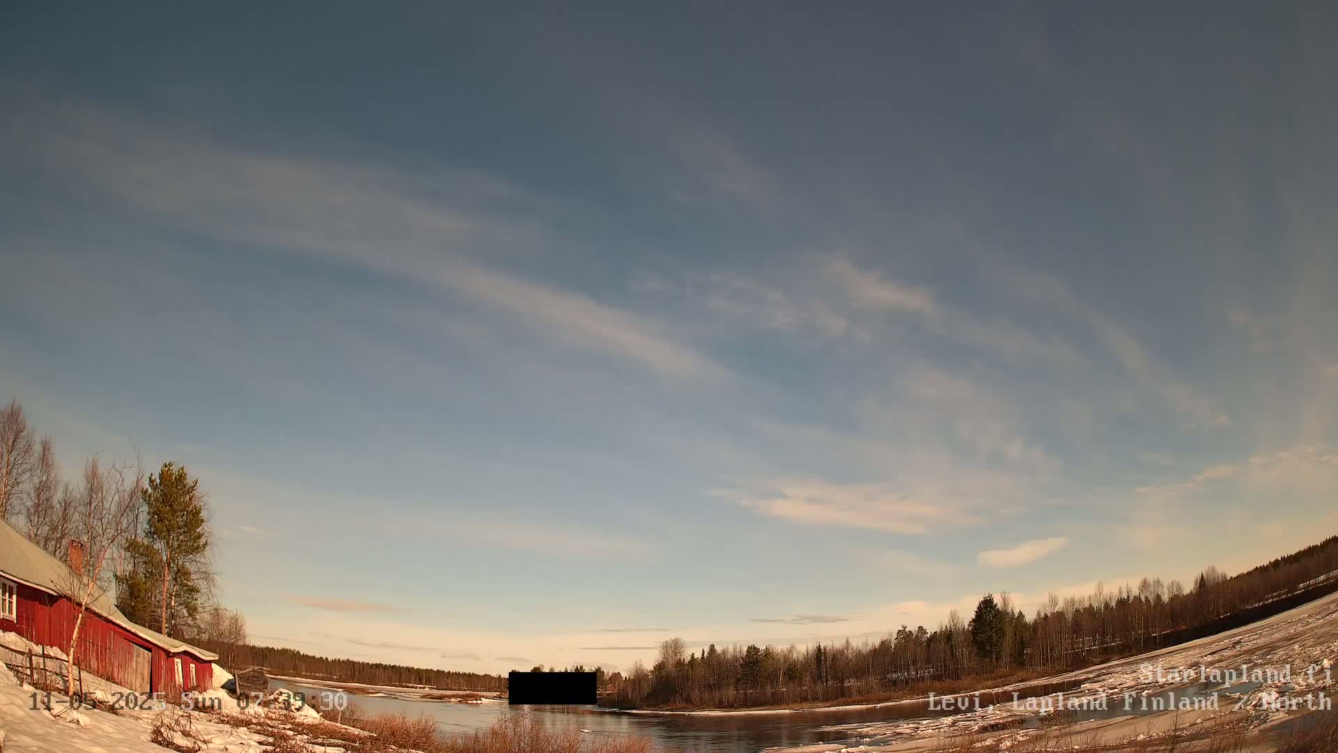 A partly cloudy sky over a snow-covered riverbank with a red building and leafless trees, under mostly clear skies.