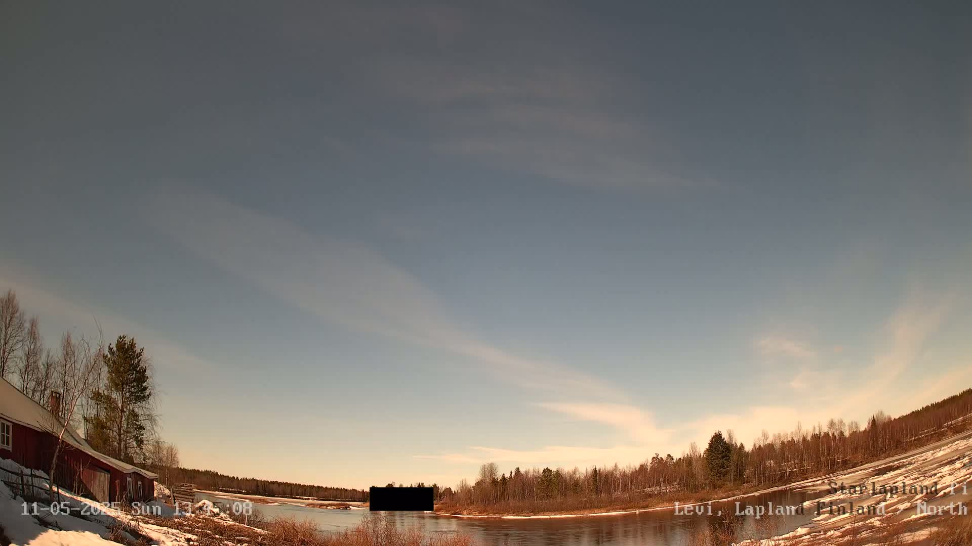 A red house sits on a snow-covered bank overlooking a calm river under a mostly clear, pale blue sky with wispy clouds.