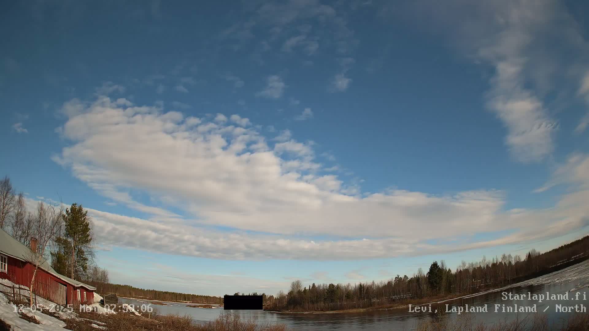 A partly cloudy blue sky overlooks a calm river bordered by trees, snow, and a red building.