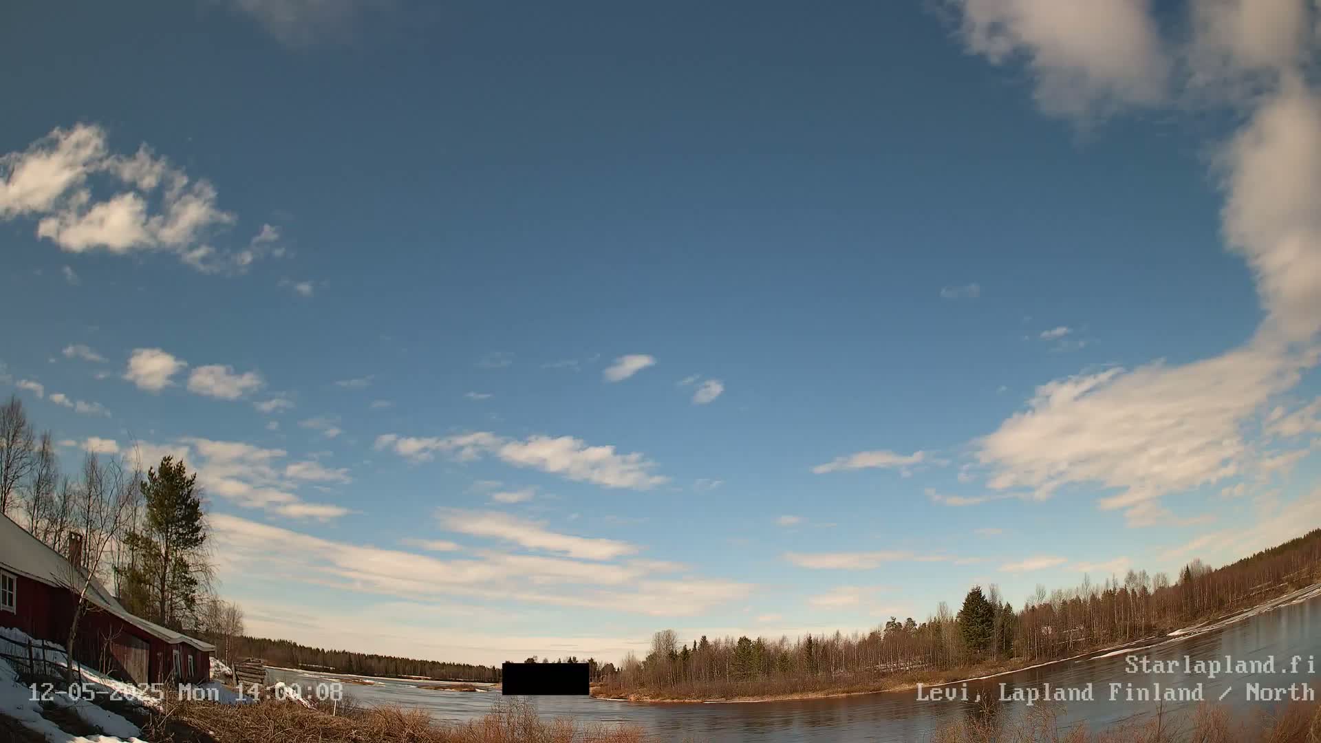 A partly cloudy sky overlooks a calm river bordered by leafless trees and a red building with some snow on the ground.