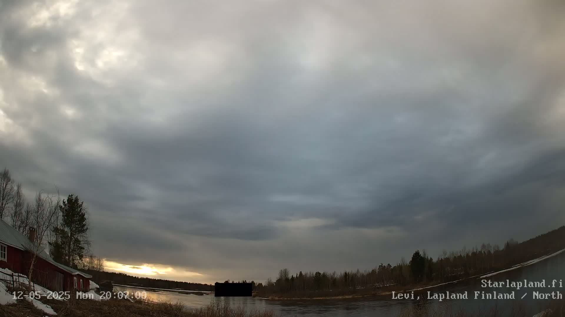 A mostly cloudy sunset over a calm river, with a red building and trees visible on the left.