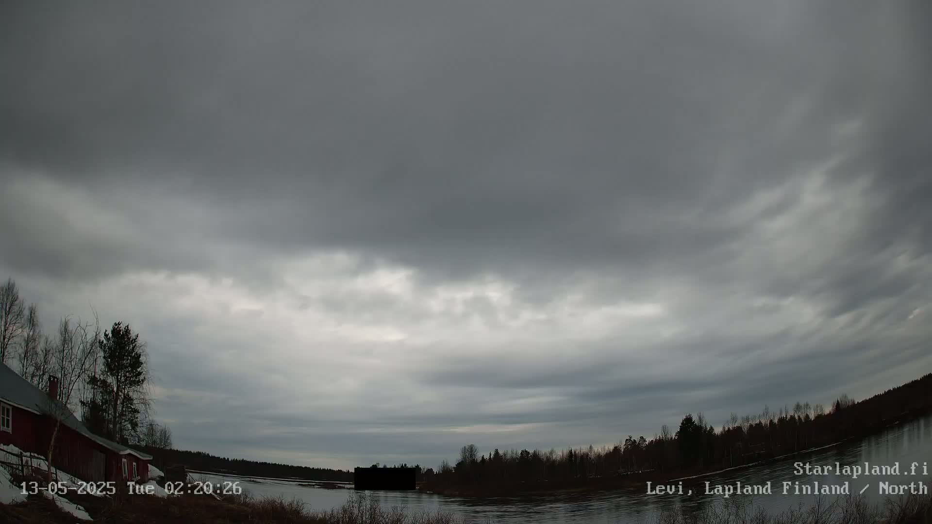 A dark, overcast sky hangs over a calm body of water bordered by leafless trees and a red house.