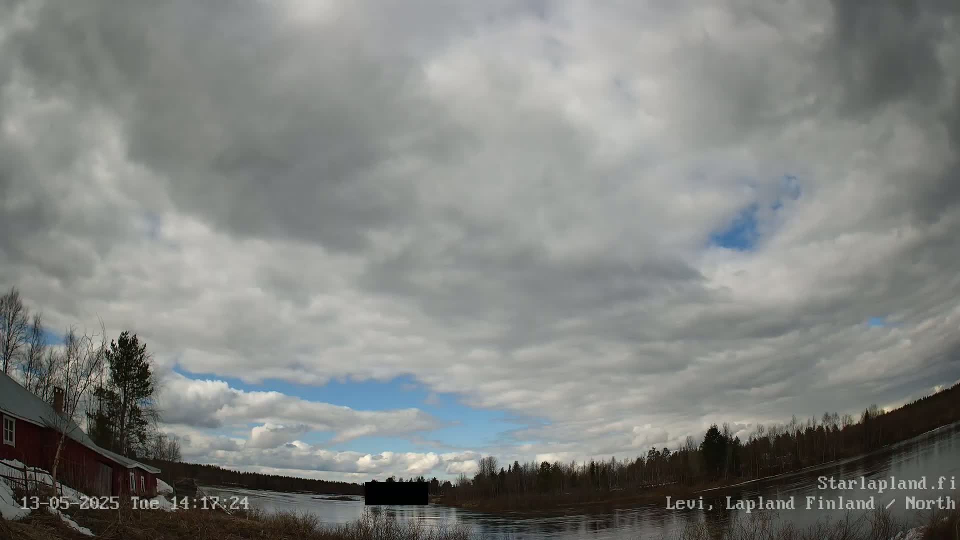 A mostly cloudy sky over a calm river, with a red building and trees visible along the riverbank.