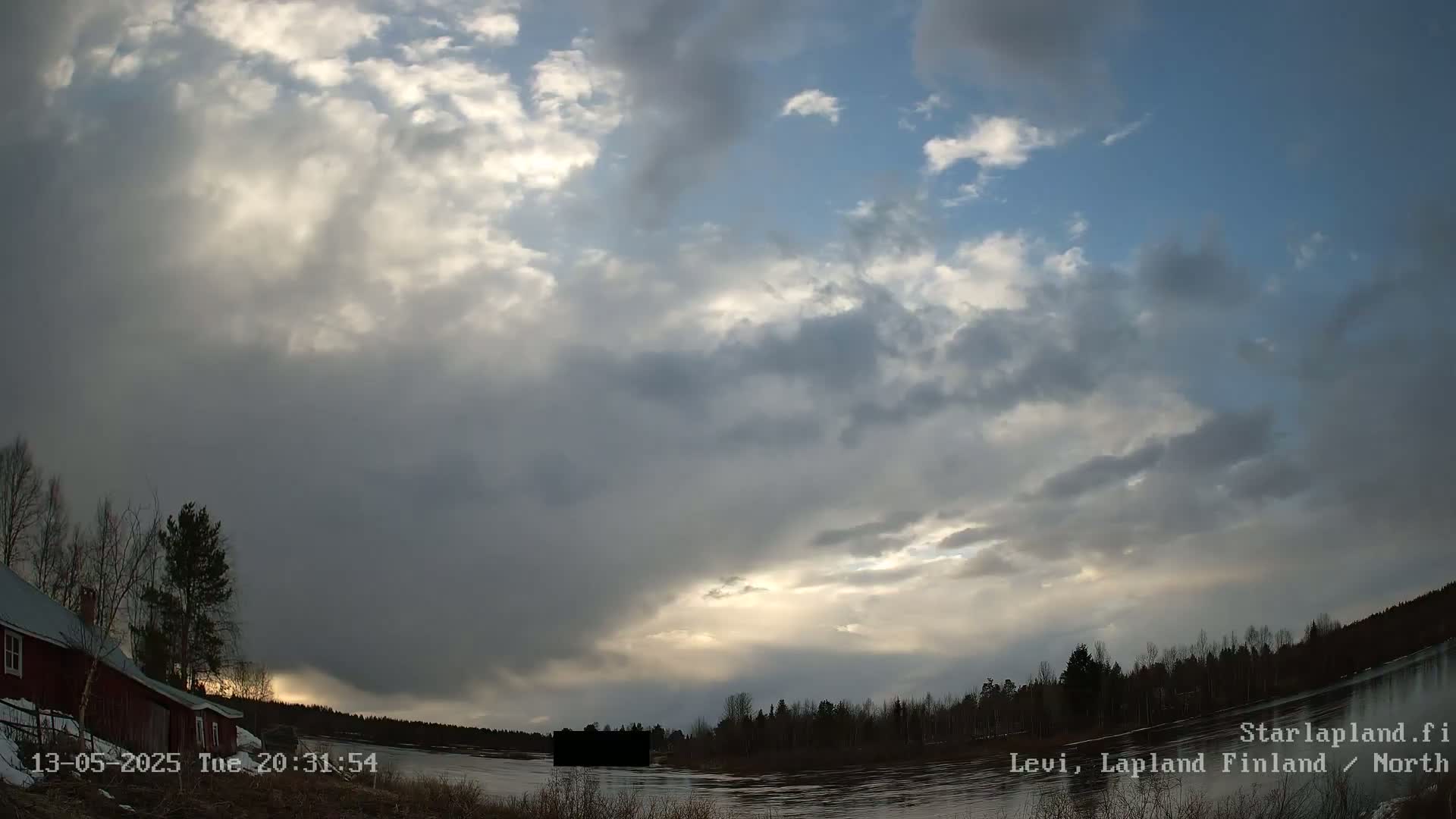 A mostly cloudy sky with some breaks of sunlight illuminates a calm body of water and a line of trees near a red building.