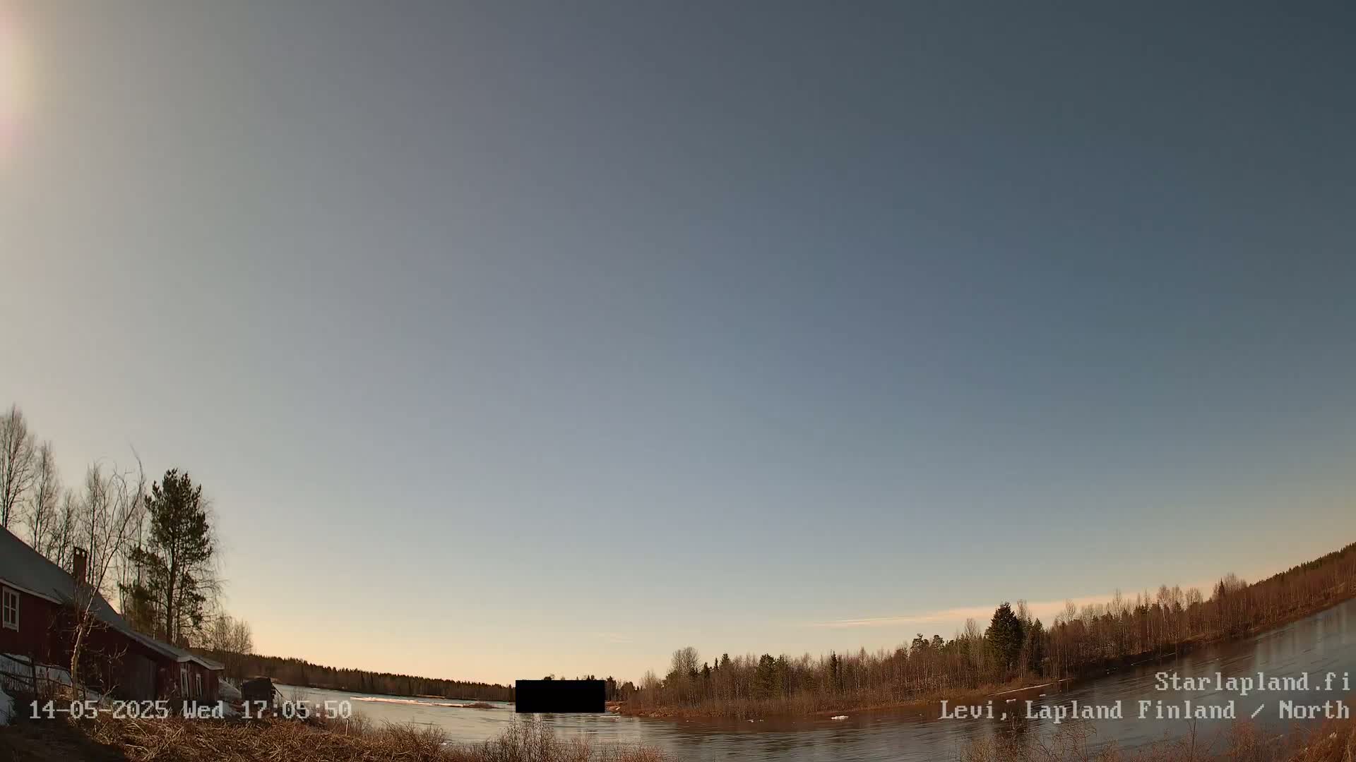 A calm body of water is seen under a mostly clear, pale blue sky, with a red building and trees visible on the left, and a shoreline of trees and shrubs on the right.