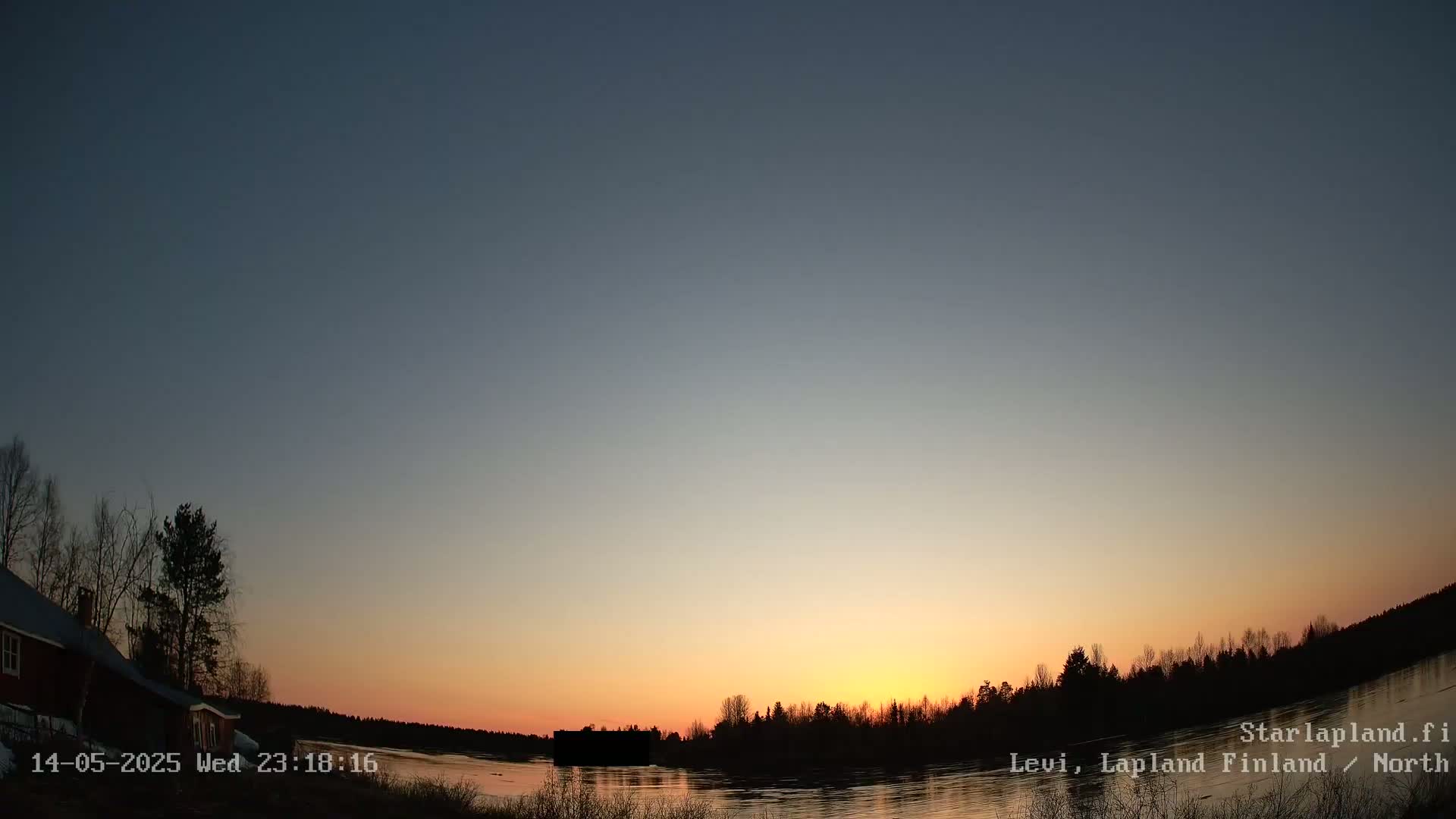 A calm lake reflects a sunset over a dark treeline, with a red building partially visible to the left.