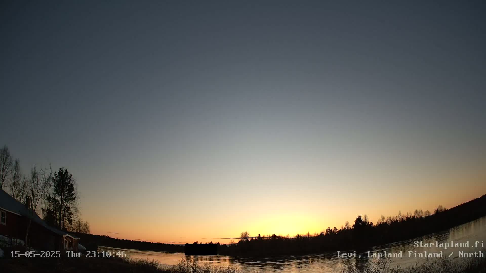 A calm lake reflects a setting sun under a clear, twilight sky, with silhouetted trees and a building on the left bank.
