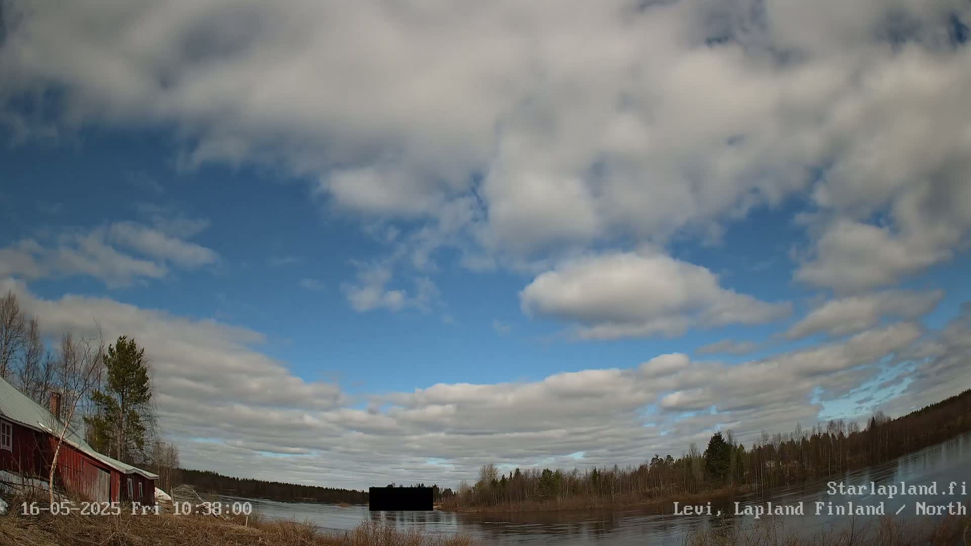 A partly cloudy sky over a calm lake, with a red building and trees visible on the left shore and a dark object in the water near the opposite shore.