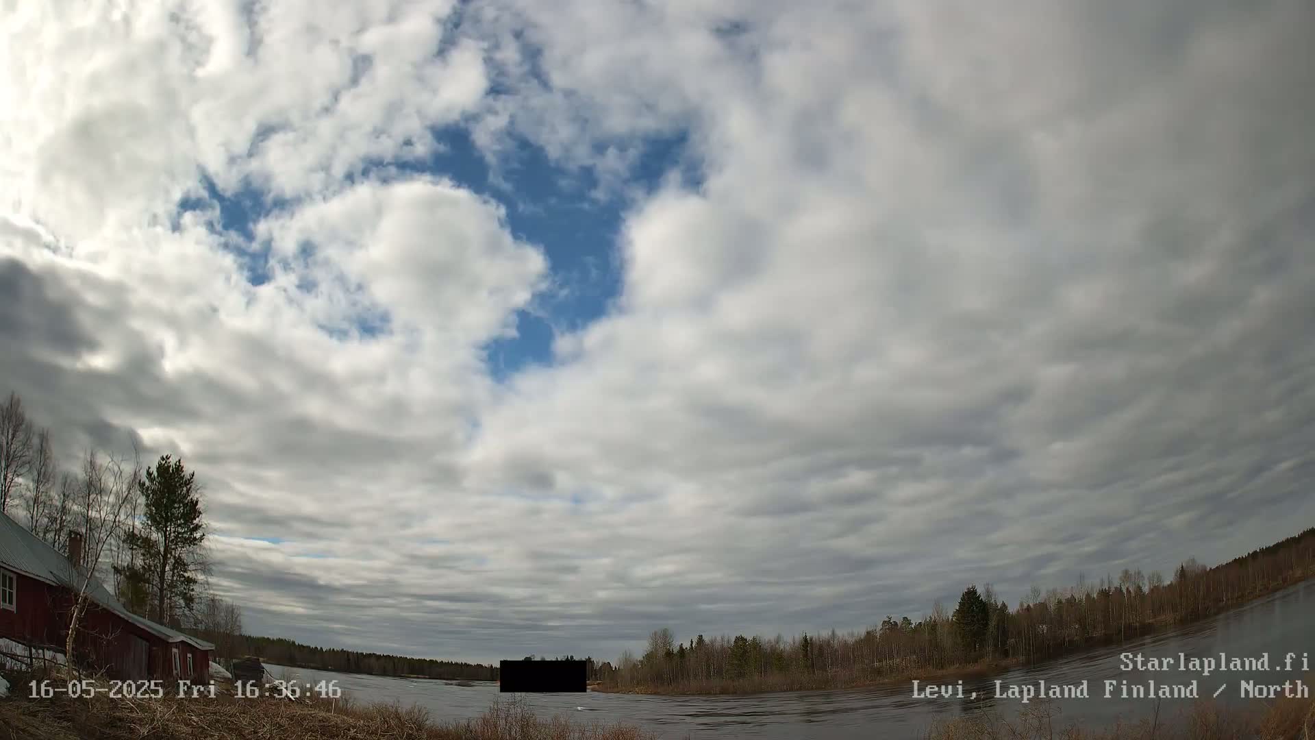 A partly cloudy sky over a calm river, with trees and a red building visible near the riverbank.