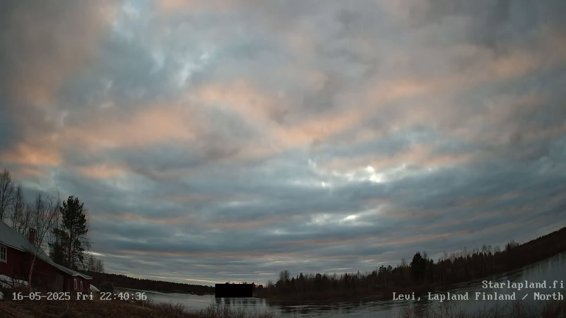 A mostly cloudy twilight sky with pale orange and gray clouds over a calm river, bordered by dark trees and a red building.