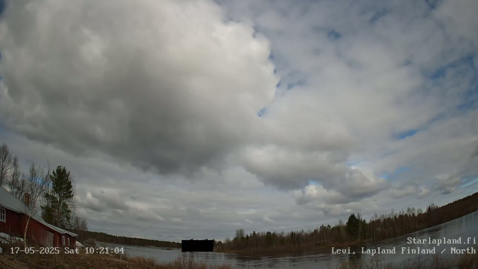 A mostly cloudy sky hangs over a calm river, with trees lining the shoreline and a red building visible on the left.