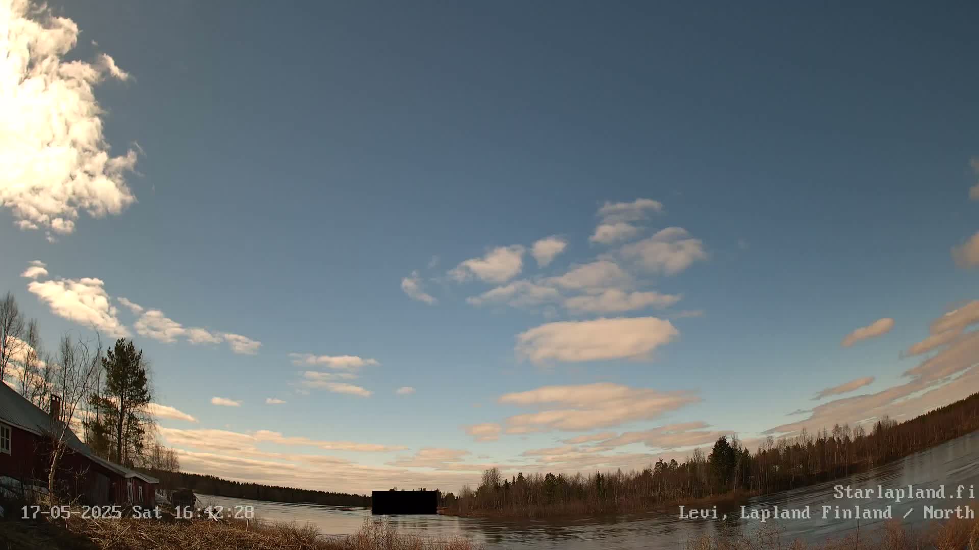 A mostly clear sky with scattered clouds over a calm lake and a shoreline with trees and a red building.