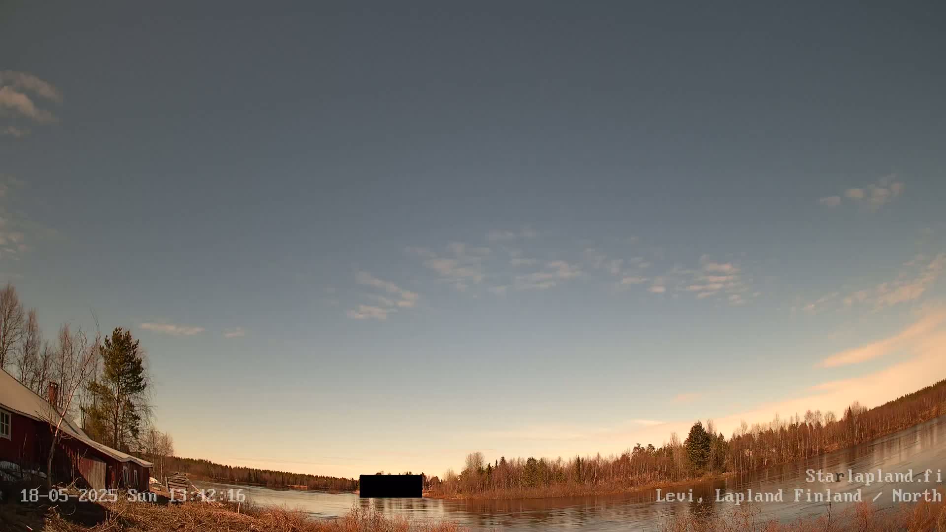 A mostly clear sky with a few wispy clouds hangs over a calm body of water, beside which sits a red house and bare trees.