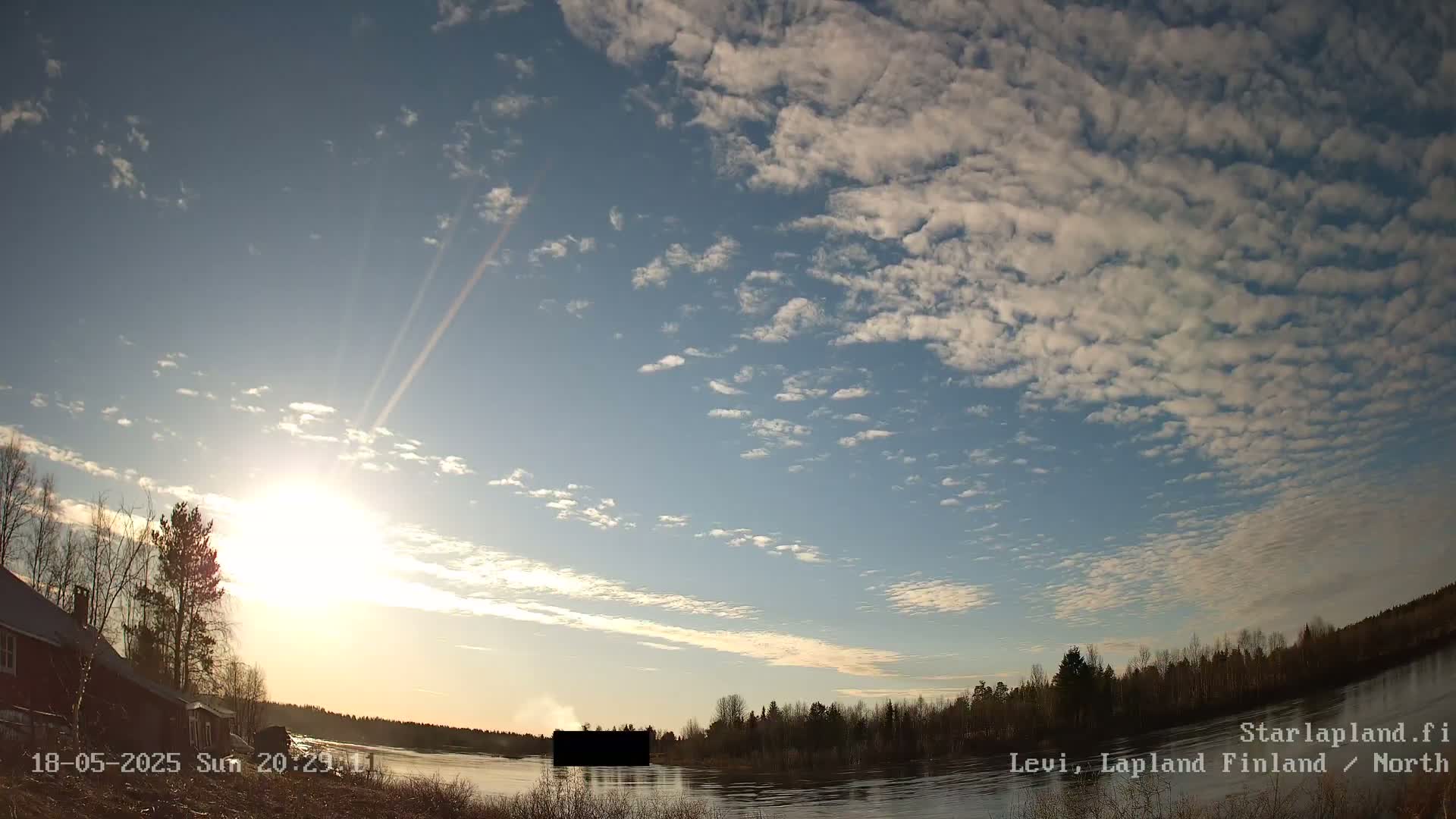 A mostly sunny day with scattered clouds over a calm river and treeline, near a building.