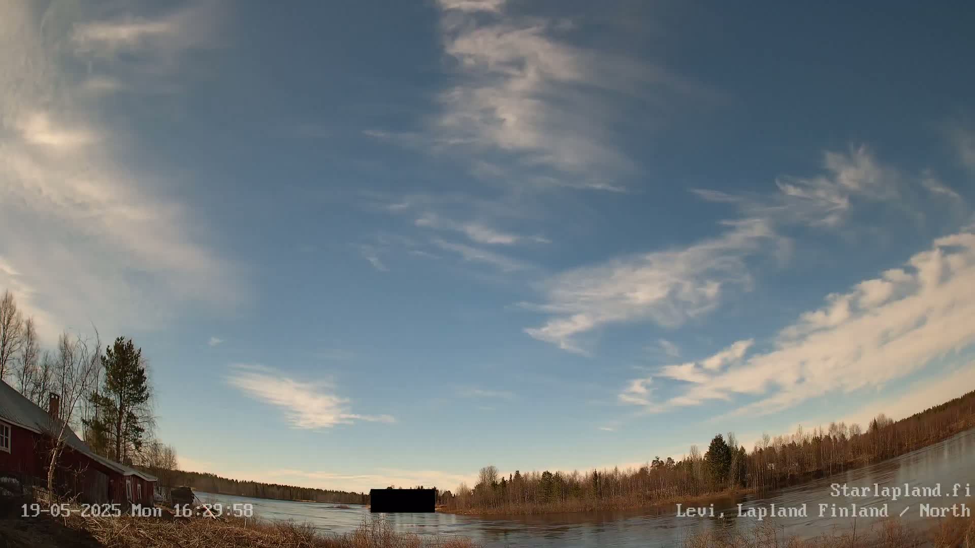 A mostly clear sky with wispy clouds over a calm river, bordered by leafless trees and a red building on the left.