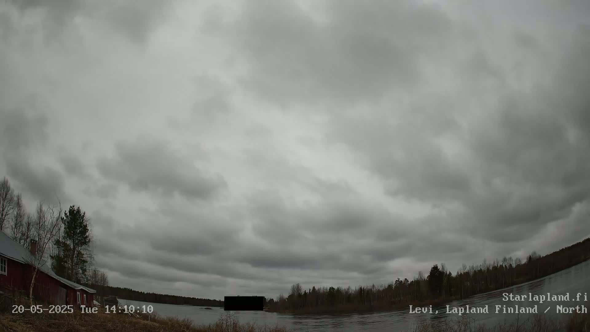 A mostly cloudy sky hangs over a calm body of water with a red building and trees visible on the left, and a treeline on the opposite shore.