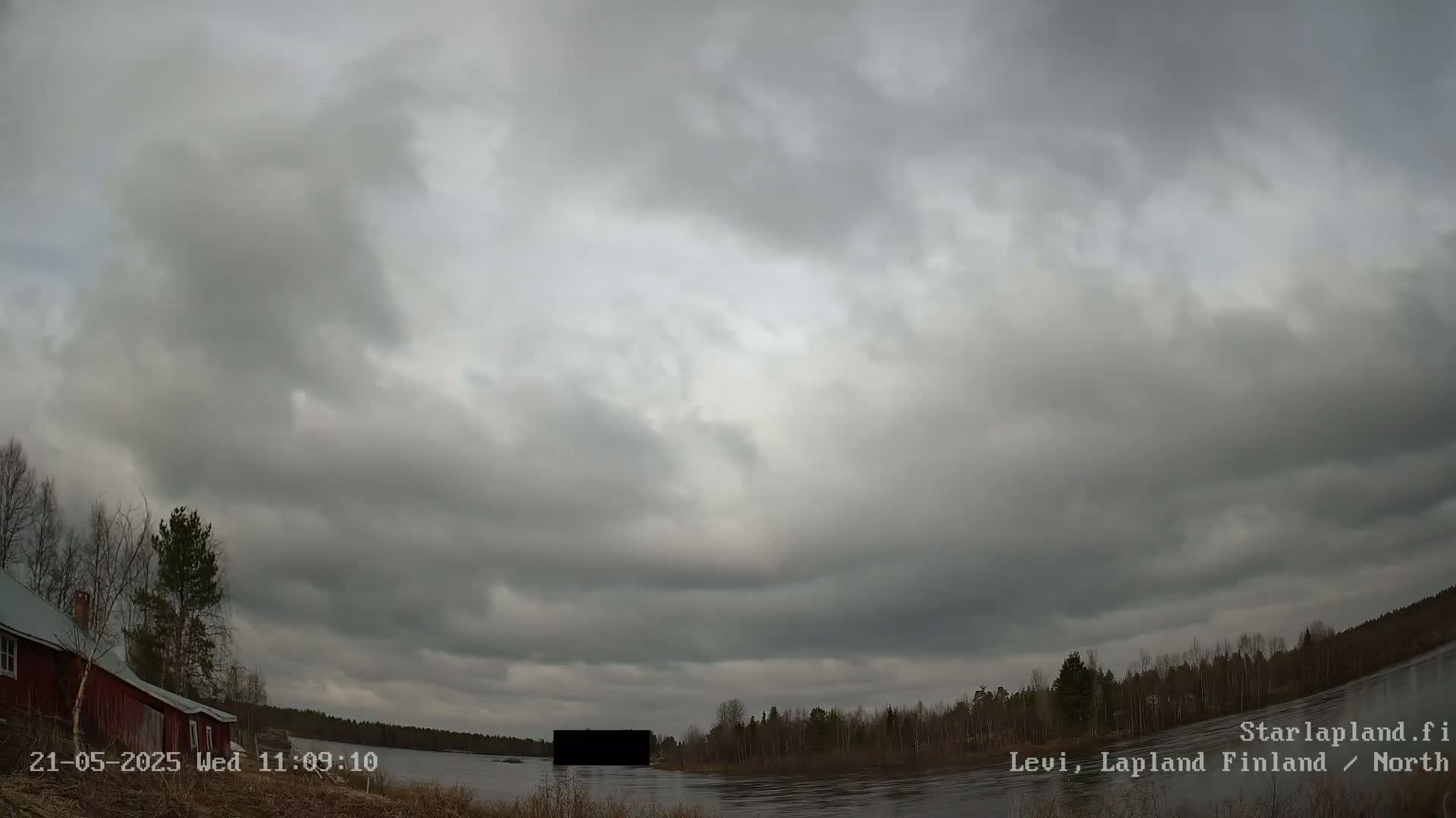A mostly cloudy sky hangs over a calm body of water with a red building and trees visible on the left, and a dark, indistinct object in the water in the center.
