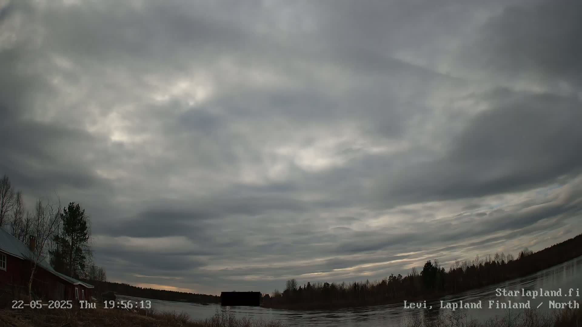 A mostly cloudy, overcast sky hangs over a calm body of water with a dark shoreline and a red building visible to the left.