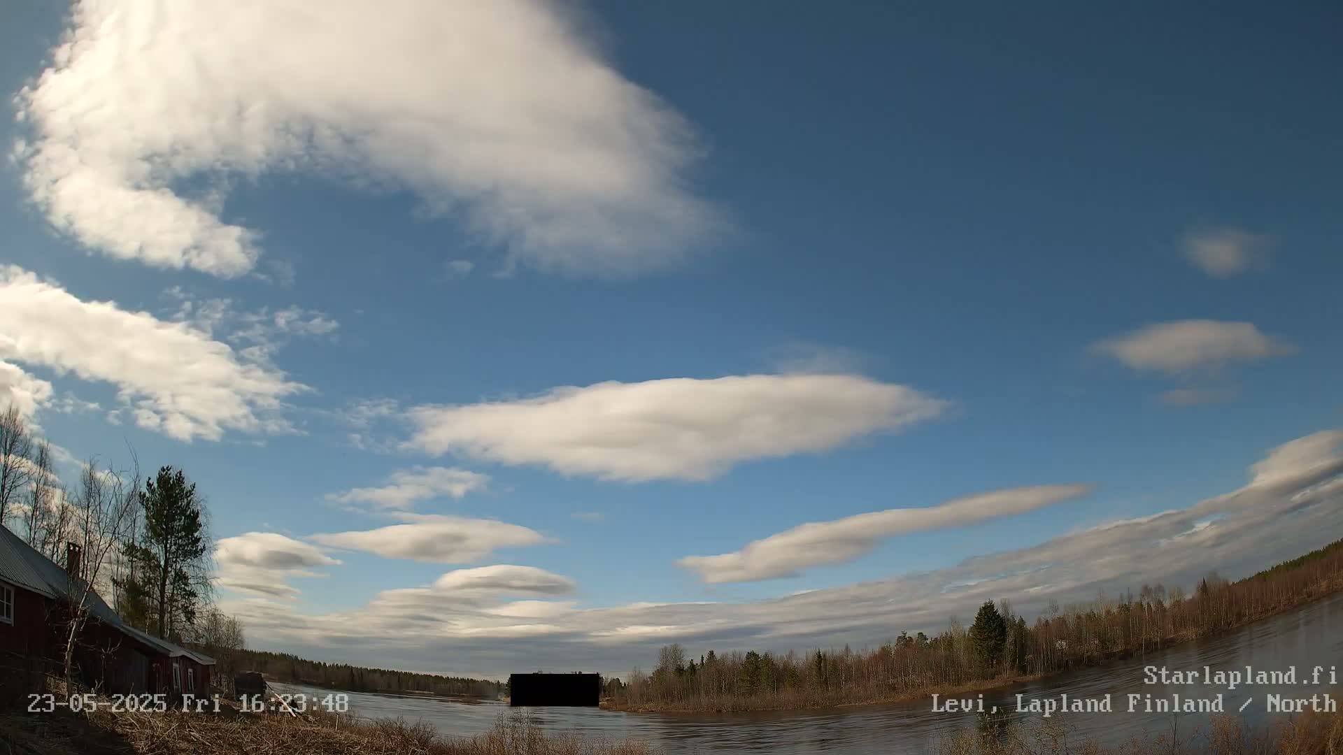 A mostly sunny day with scattered lenticular clouds over a calm river and sparsely wooded shoreline with a red building visible on the left.